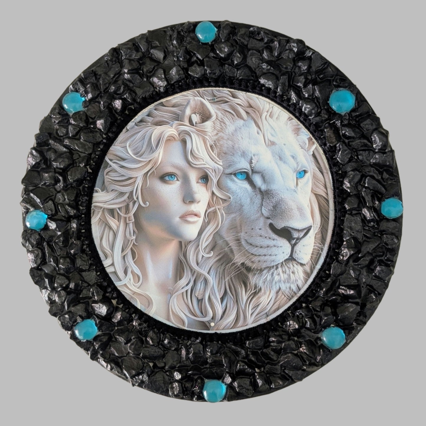 "Blue Mystique" is a handmade canvas featuring a print of a white lion and woman with striking blue eyes, framed by black and bright blue stones.