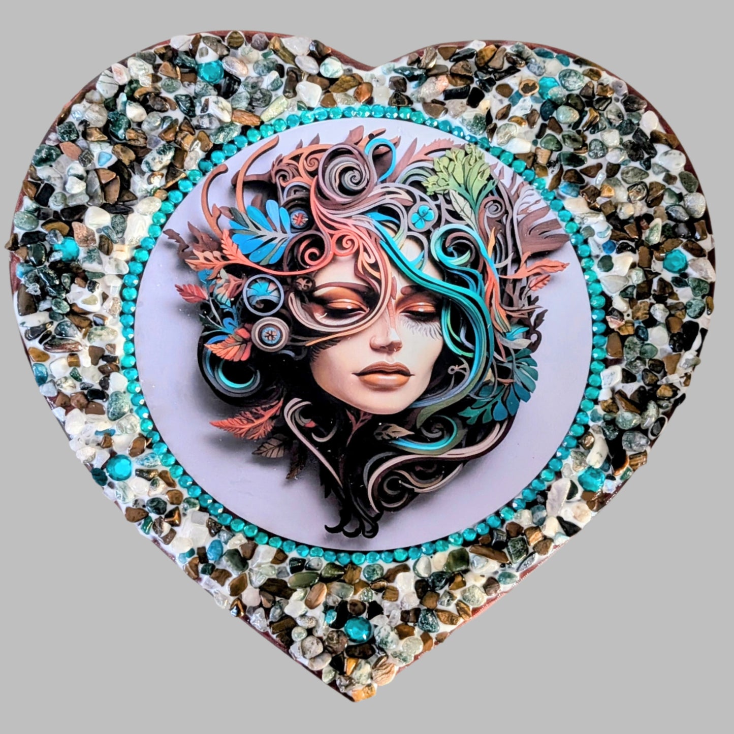 "Echoes of the Heart" is a handmade heart shaped canvas with a print of a woman surrounded by swirling blue and earthy coloured leaves, decorated with earthy coloured stones & pebbles.
