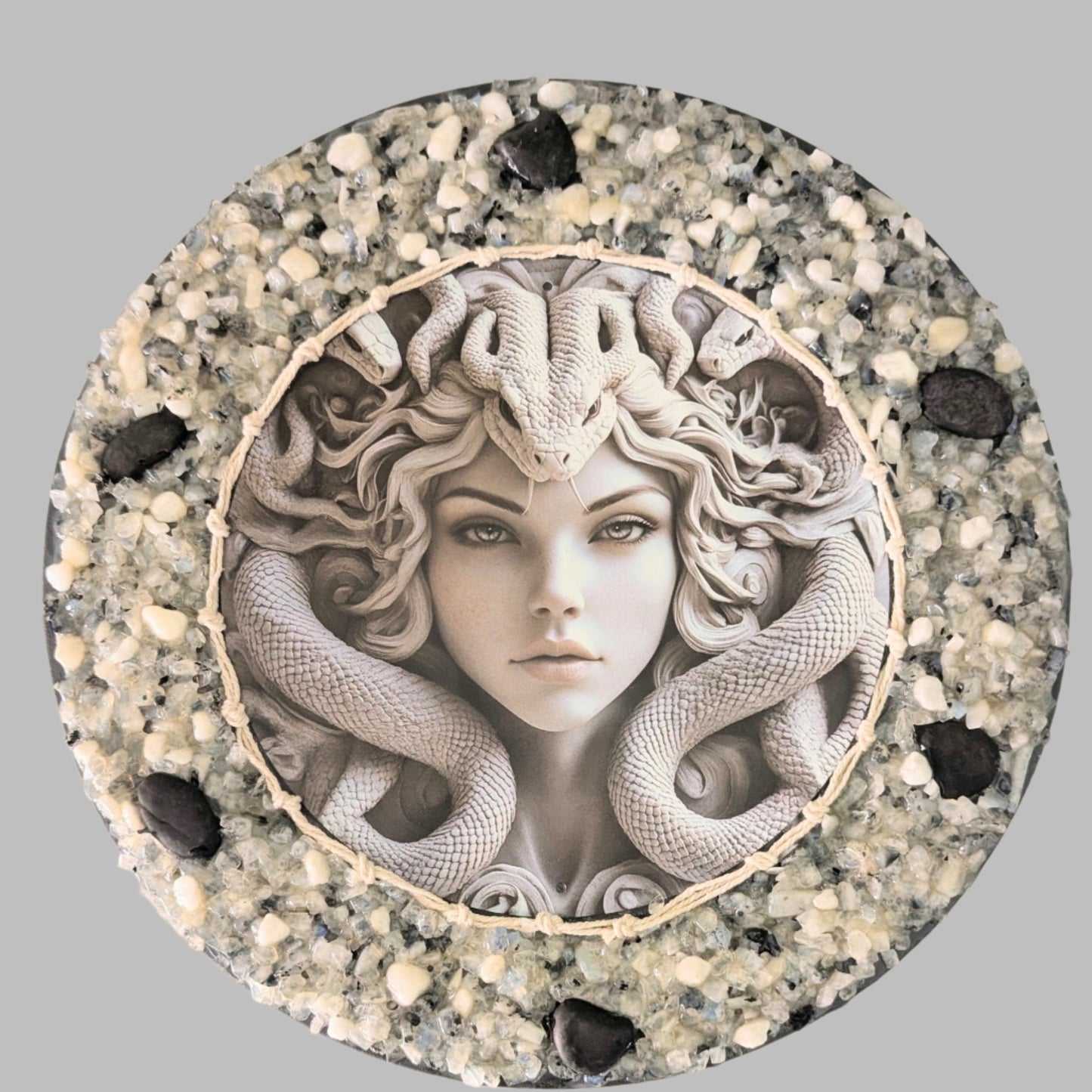"The Serpent's Glare" is a handmade canvas featuring a print of Medusa with striking eyes, framed by snakes and decorated with bold black stones and white and clear stones & pebbles.