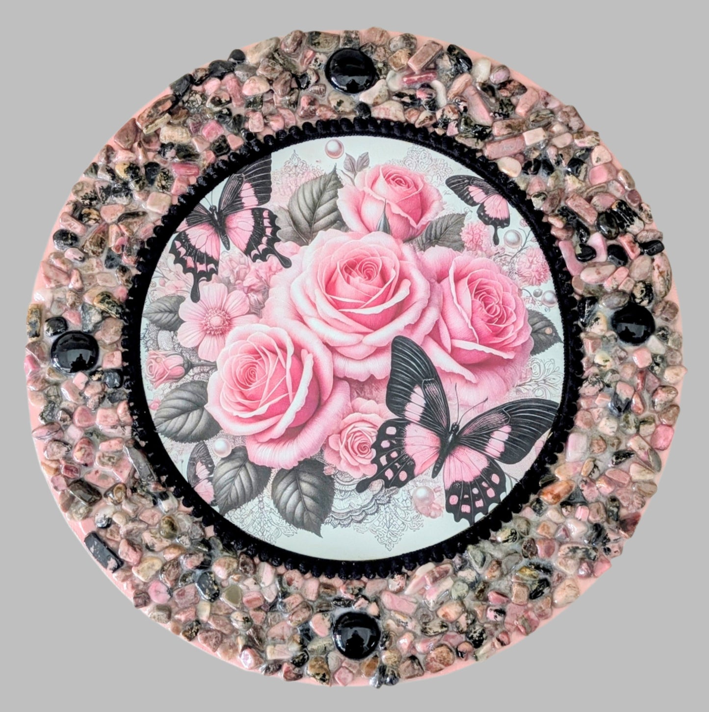 "Rose-Tinted Wings" is a handmade canvas featuring a print of a pink butterfly and roses, accented with black stones and pink and earthy coloured pebbles, symbolising transformation and beauty.