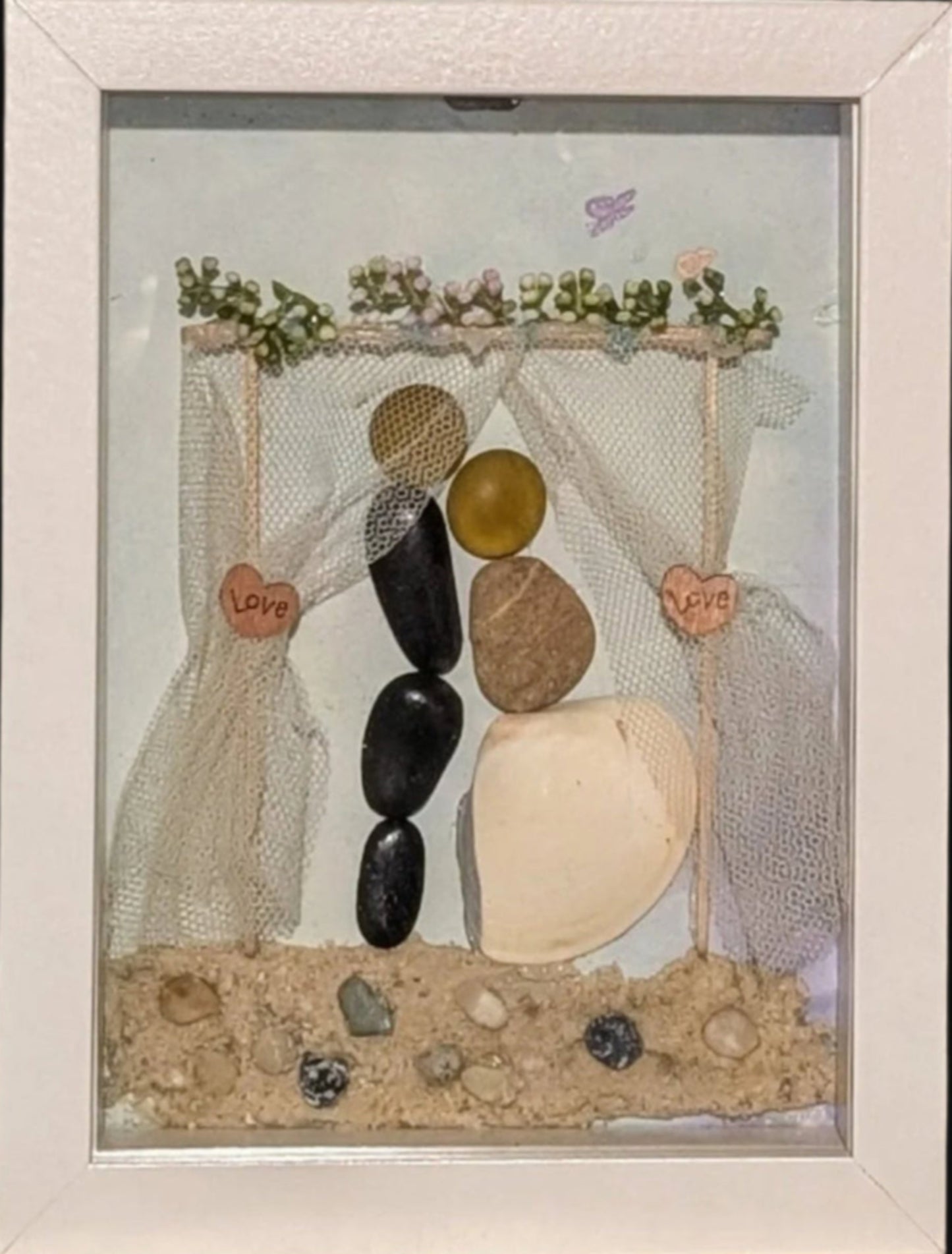 Framed stone art of a couple exchanging vows on the beach, with a seashell-adorned gown, love heart arch, and romantic pink flowers celebrating a seaside wedding.