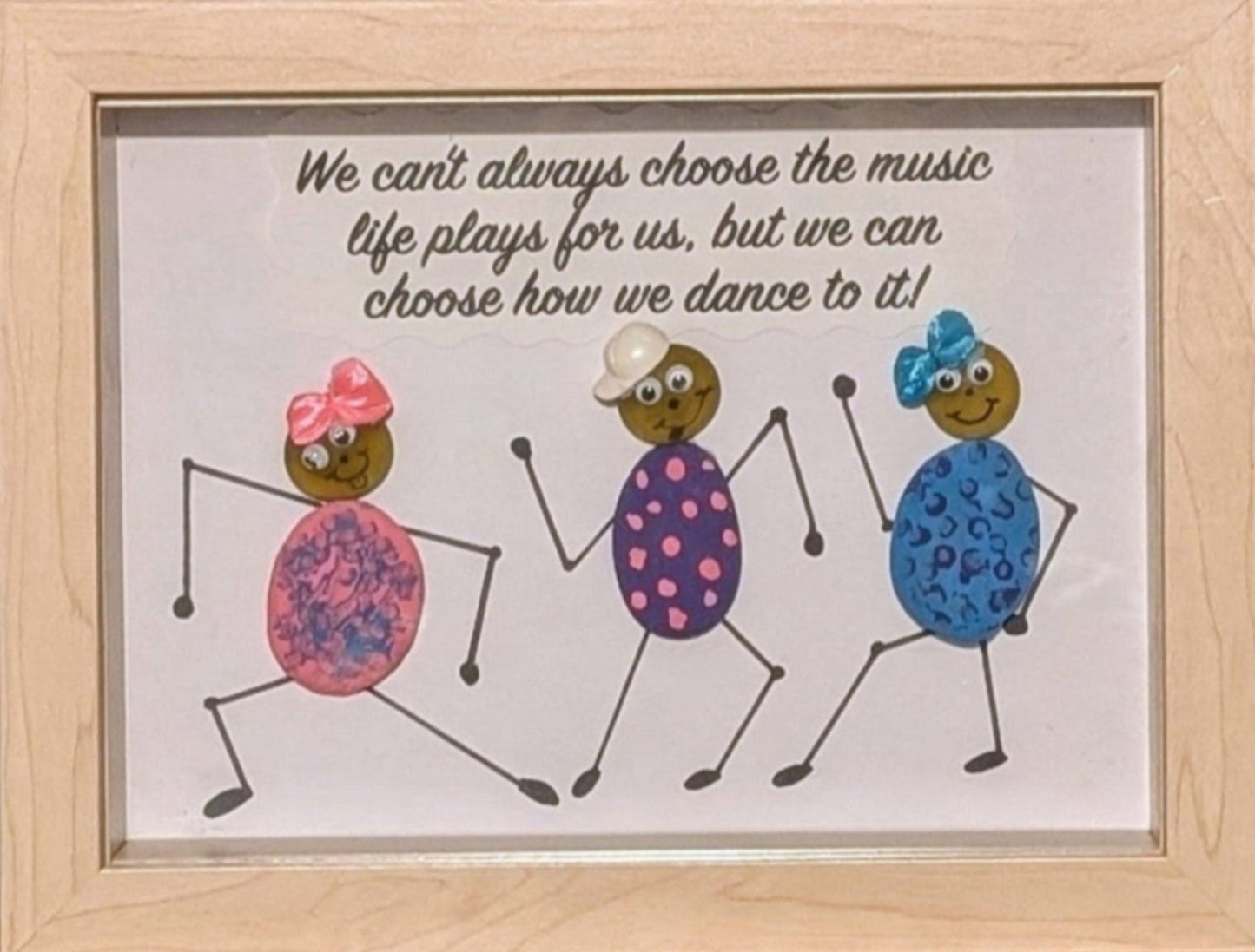 Framed pebble art of three dancing figures adorned in bright colours and bows, symbolizing joy, freedom, and the connection of unity in celebration.