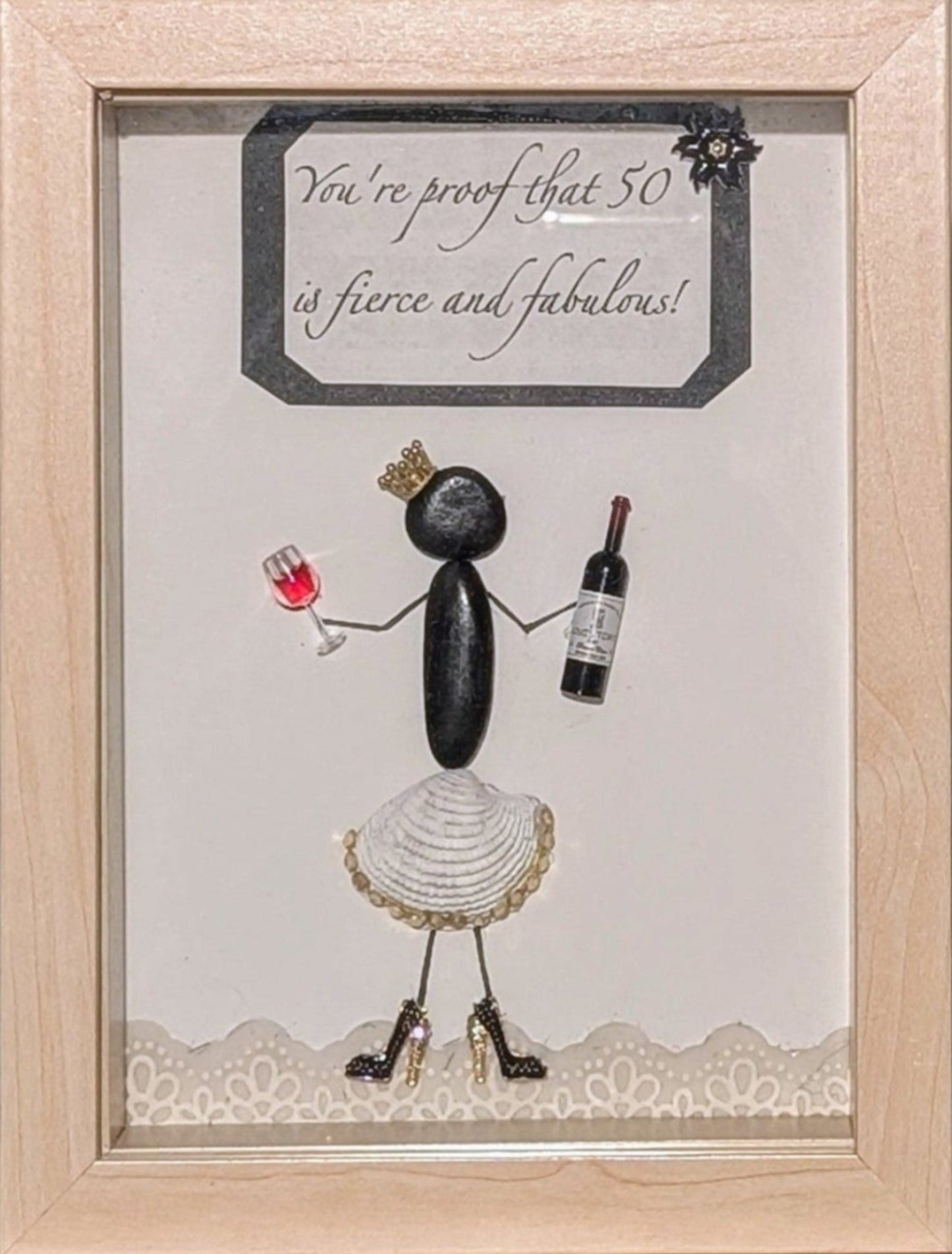 Framed pebble art of a poised woman with a golden crown, wine bottle, and glass, in a white shell skirt and black stilettos, symbolizing celebration and elegance.