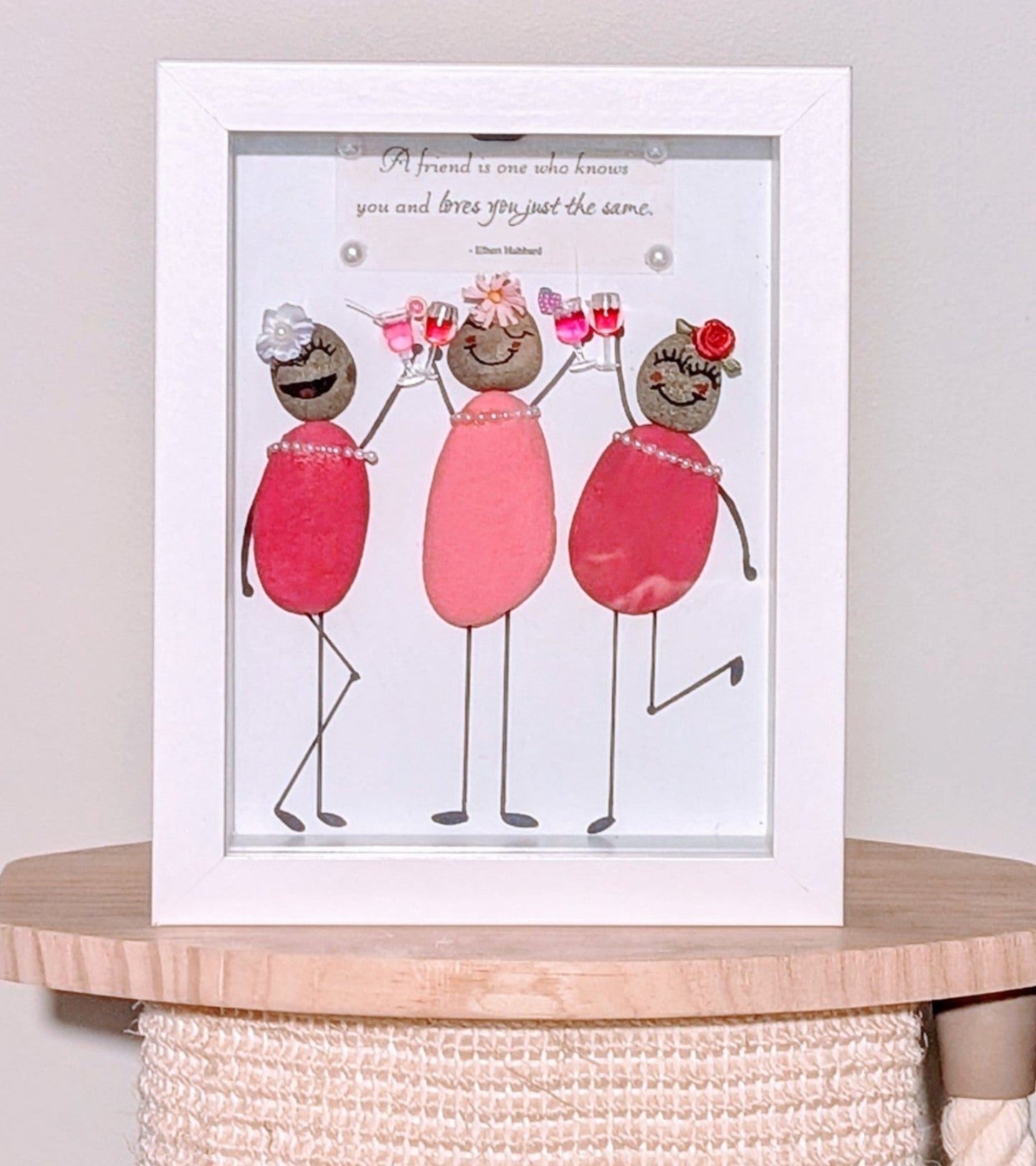 Framed pebble art of three women raising a toast with cocktail glasses, wearing pink dresses with pearls and flower headpieces, symbolising friendship and celebration.