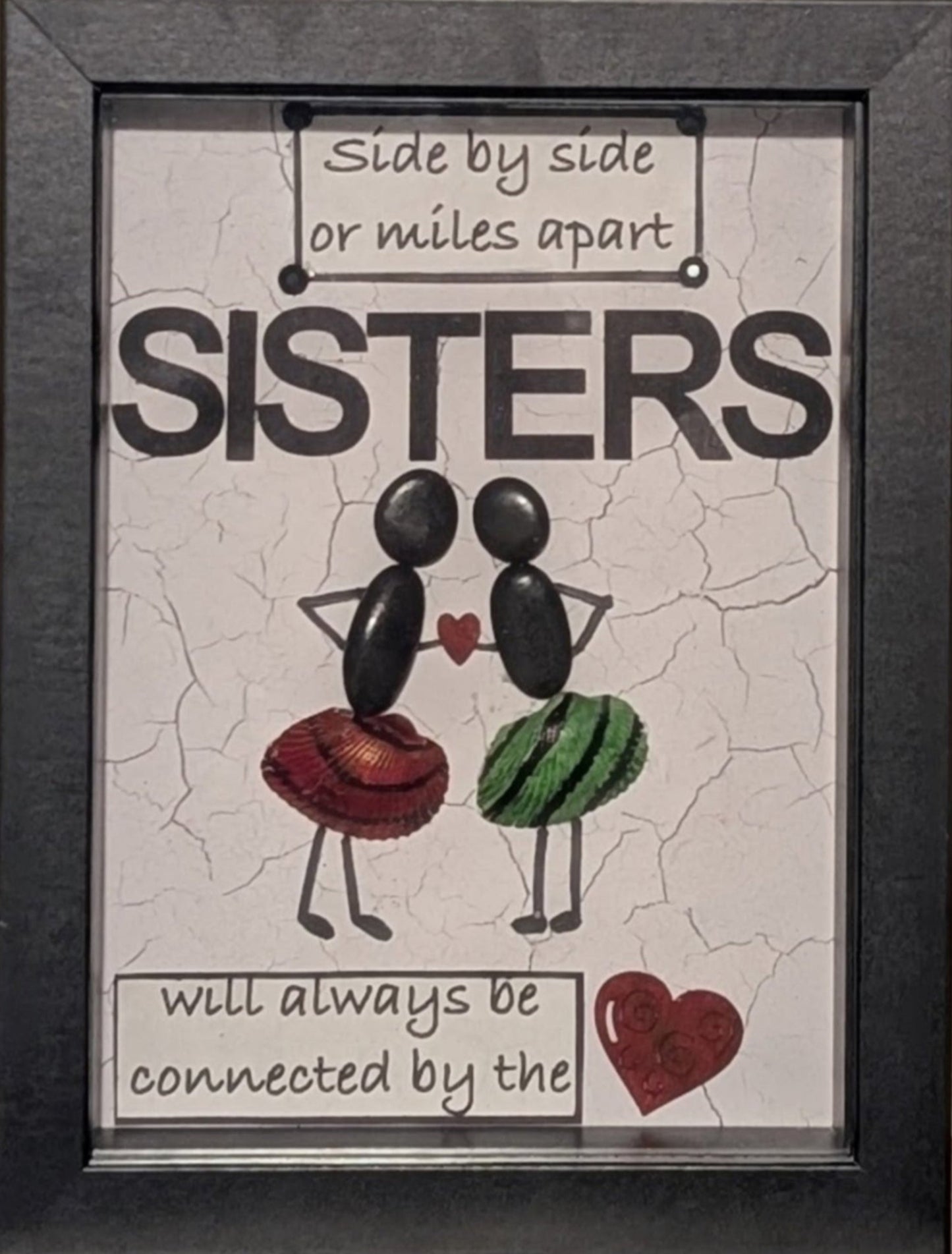Framed pebble art of two sisters holding a heart between them, with uniquely painted red, black, and green shells on their skirts, symbolising unity and love.