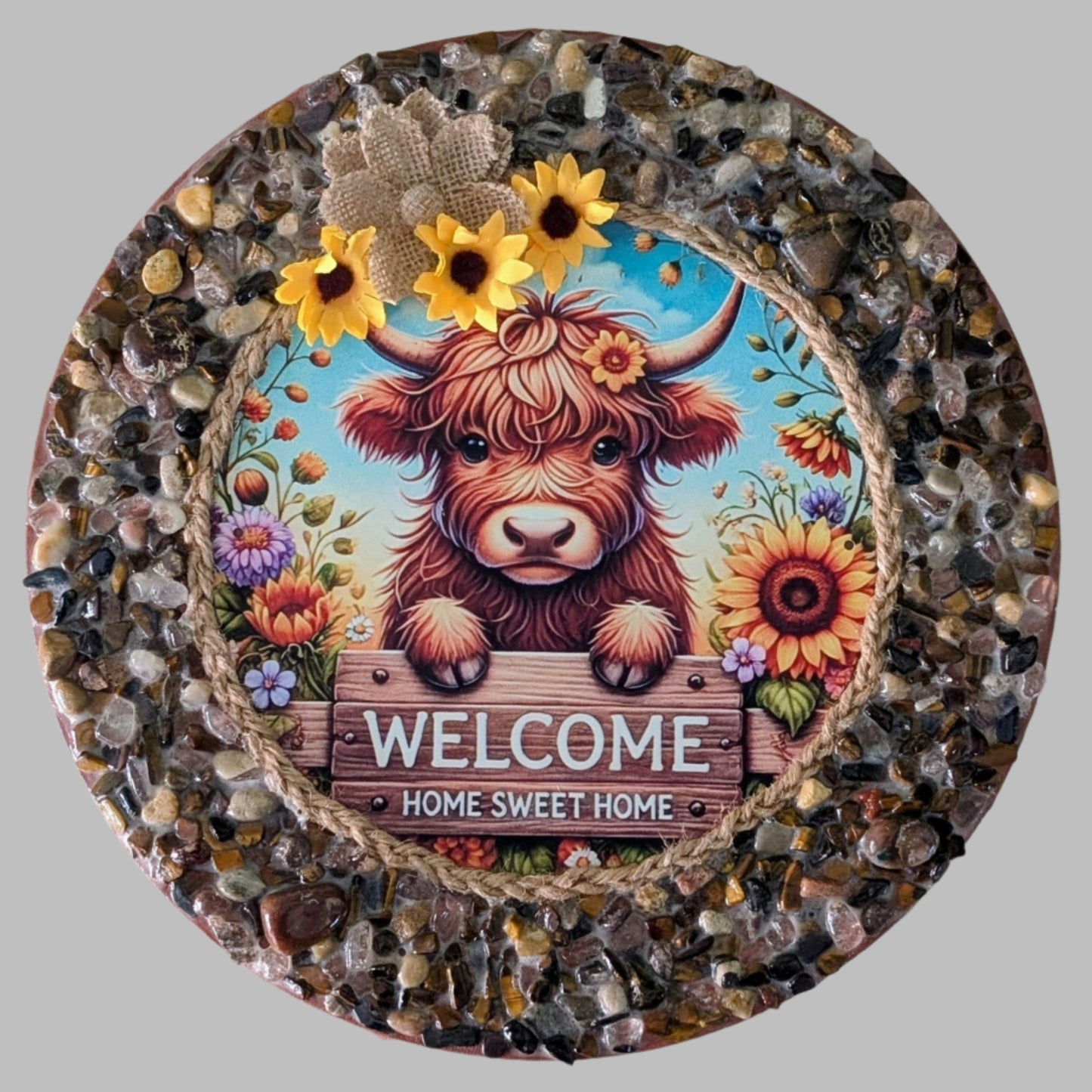 "Sweet Cow-nection" is a handmade canvas featuring a print of a cow with a "Welcome Home" sign, surrounded by colorful flowers and decorated with earthy stones & pebbles.