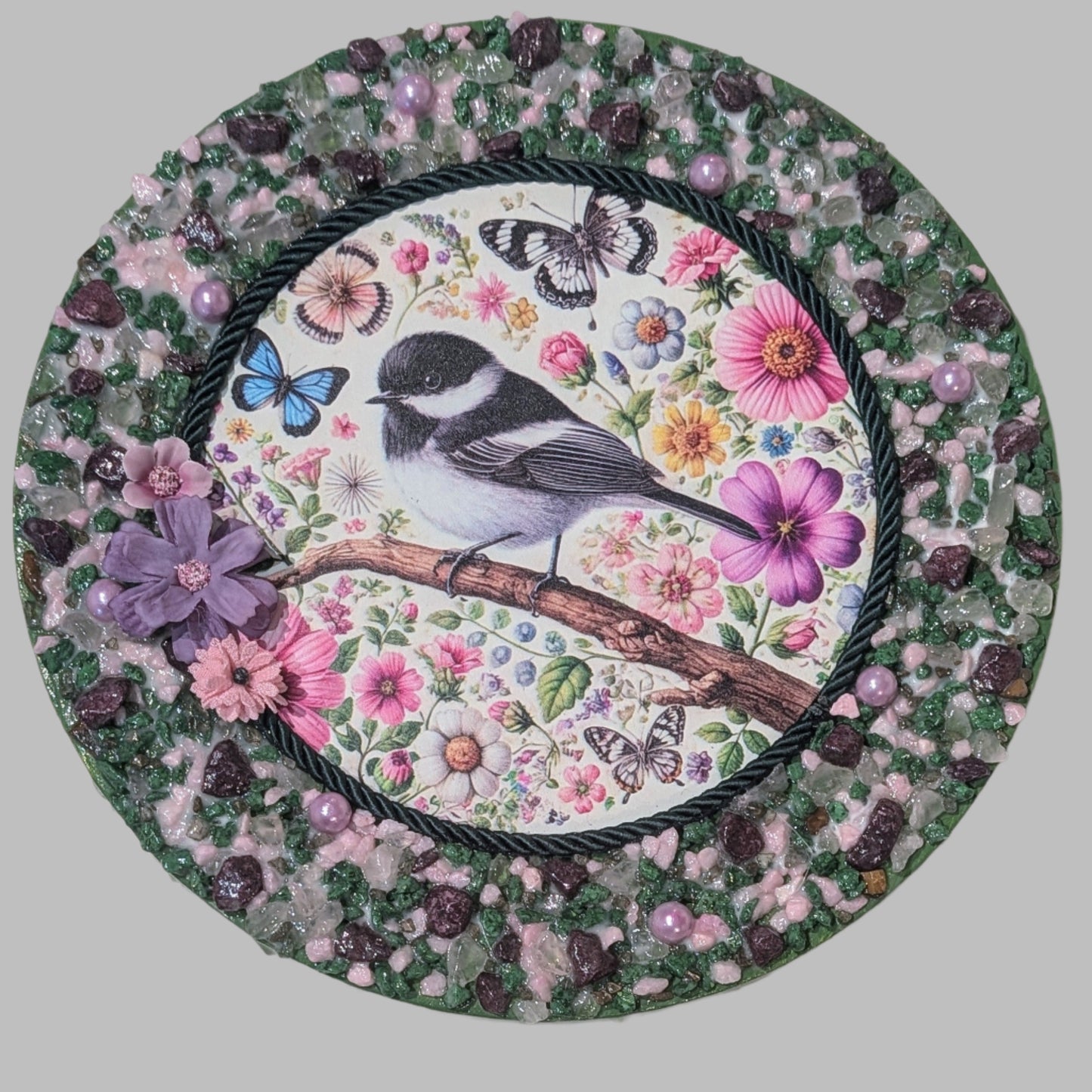 Handmade canvas featuring a print with a black and white bird on a branch, surrounded by colourful flowers, butterflies, green rope, lilac pearls, and pink/purple flowers.