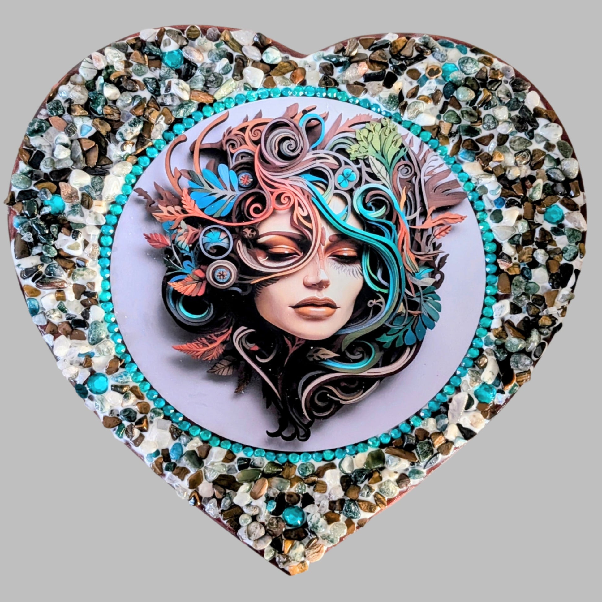 "Echoes of the Heart" is a handmade heart shaped canvas with a print of a woman surrounded by swirling blue and earthy coloured leaves, decorated with earthy coloured stones & pebbles.