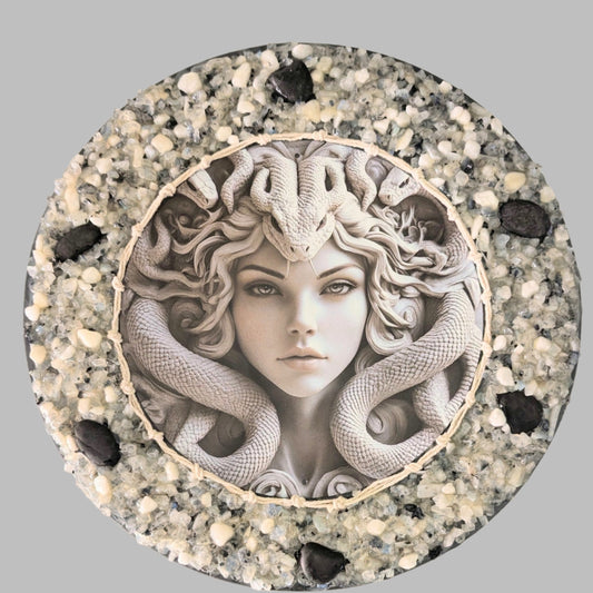 "The Serpent's Glare" is a handmade canvas featuring a print of Medusa with striking eyes, framed by snakes and decorated with bold black stones and white and clear stones & pebbles.