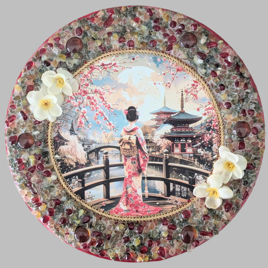 "The Geisha's Reflection" is a handmade canvas featuring a print of a geisha on a bridge, blooming trees, and reflective water, accented with red and earthy stones and cream flowers.