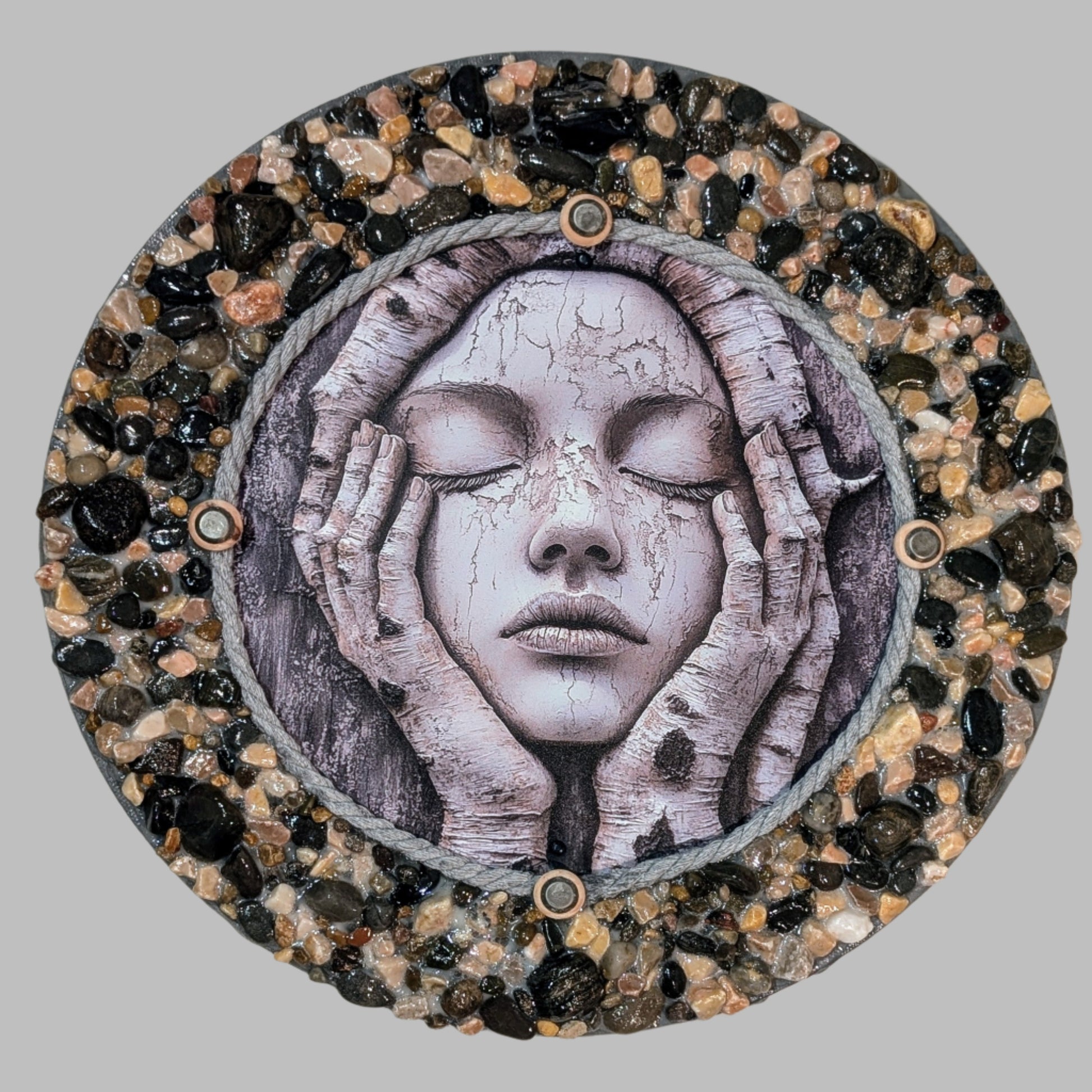 Handmade canvas with a print of a woman holding her face, surrounded by tree branches, decorated with grey rope, metal studs, and earthy stones and pebbles.