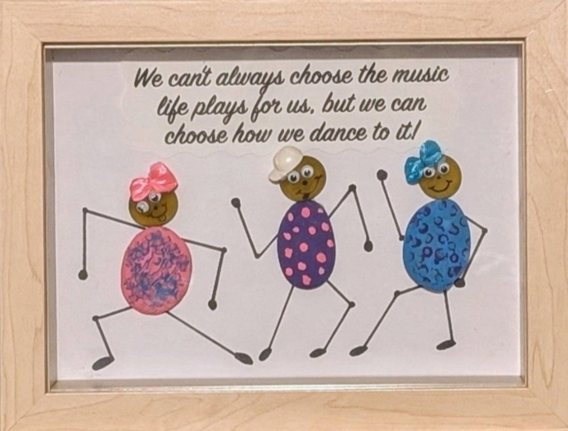 Framed pebble art of three dancing figures adorned in bright colours and bows, symbolizing joy, freedom, and the connection of unity in celebration.