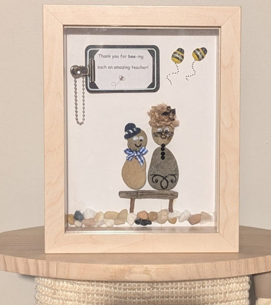 Framed pebble art of a teacher and student in a classroom, with buzzing bees and a clipboard, symbolising the joy of learning and the energy of education.