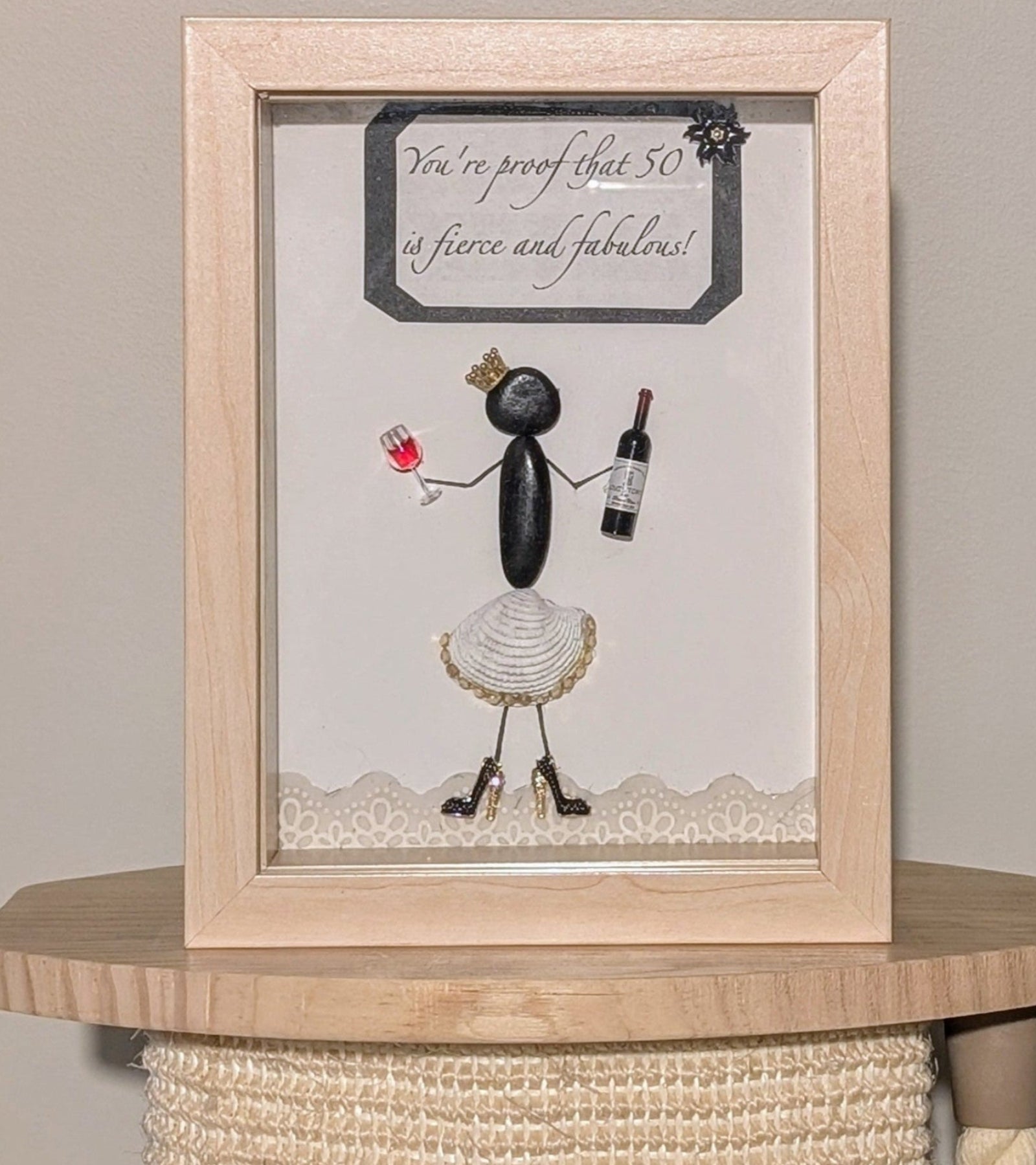 Framed pebble art of a poised woman with a golden crown, wine bottle, and glass, in a white shell skirt and black stilettos, symbolizing celebration and elegance.