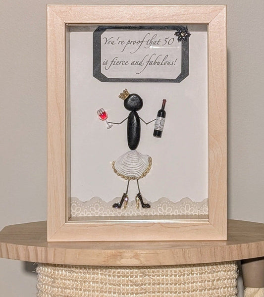Framed pebble art of a poised woman with a golden crown, wine bottle, and glass, in a white shell skirt and black stilettos, symbolizing celebration and elegance.
