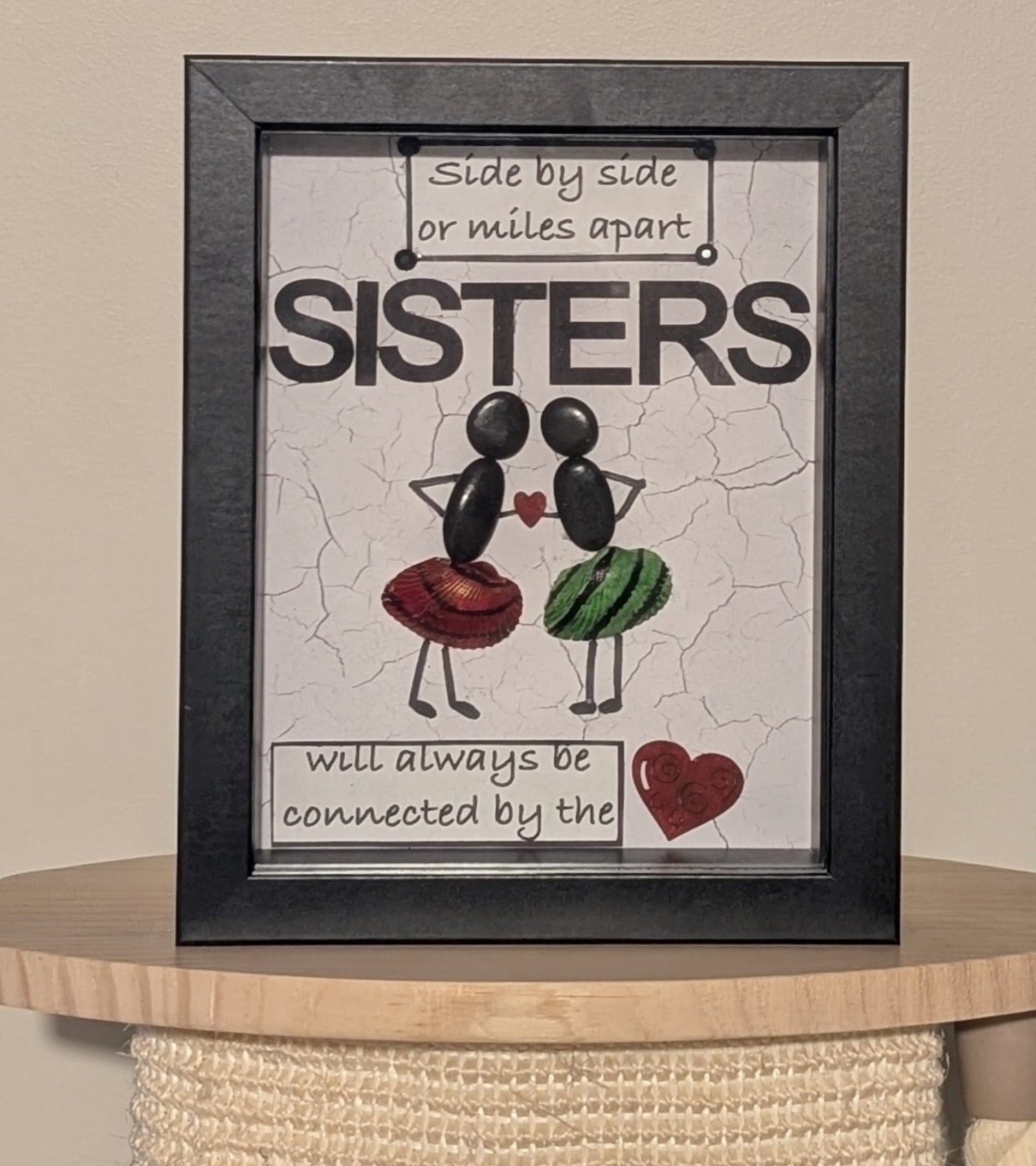 Framed pebble art of two sisters holding a heart between them, with uniquely painted red, black, and green shells on their skirts, symbolising unity and love.