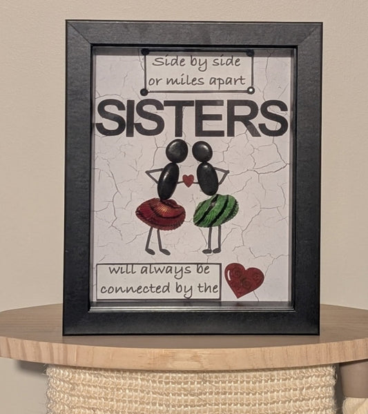 Framed pebble art of two sisters holding a heart between them, with uniquely painted red, black, and green shells on their skirts, symbolising unity and love.